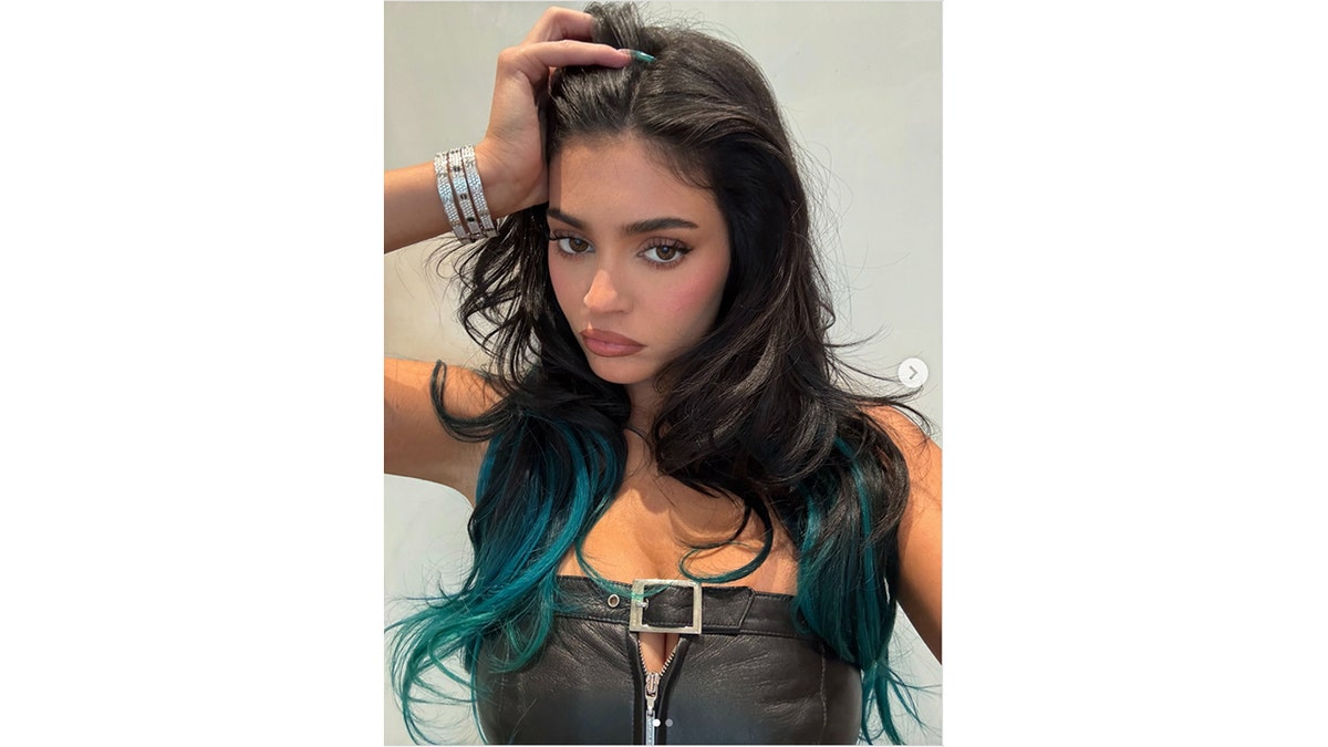 Kylie Jenner poses with her hand in her hair, showing black hair with teal ends while wearing a black leather zip-up top.