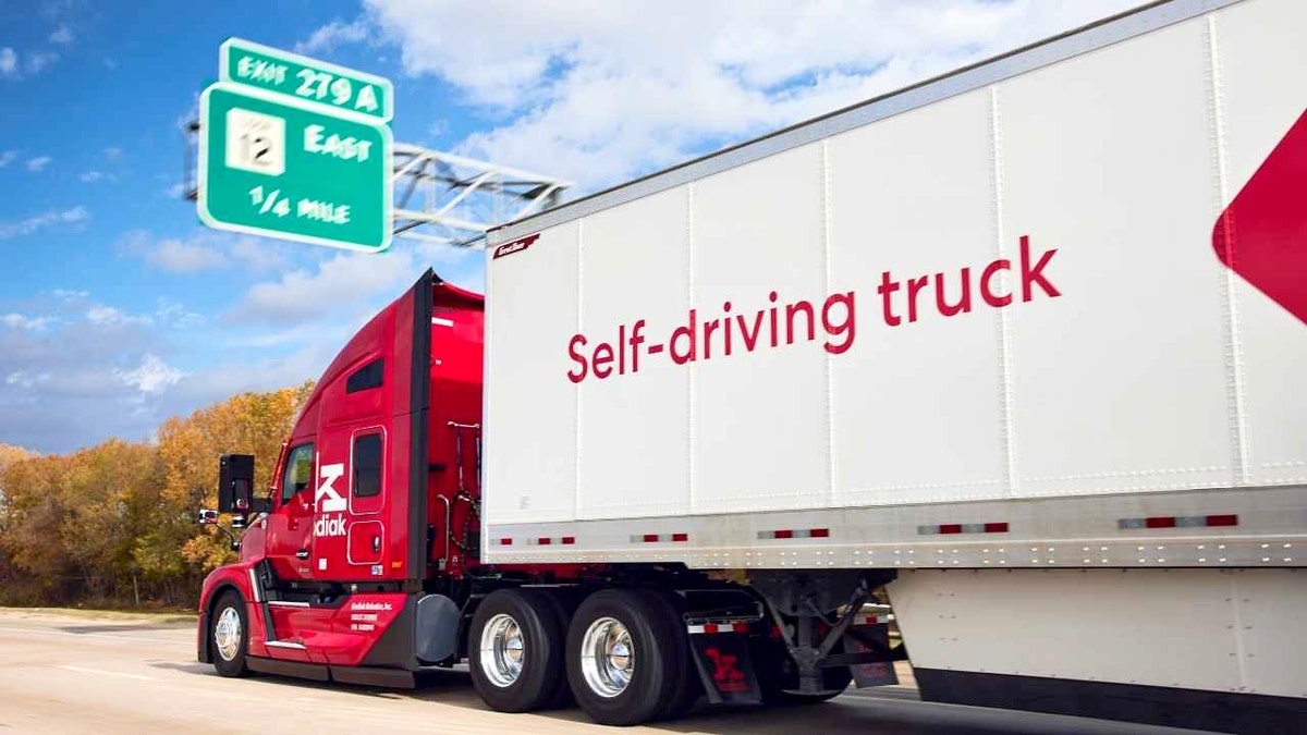 Kodiak autonomous truck system ties top safety score with human fleets 2 A red Kodiak self-driving semi-truck drives on a highway under a blue sky near an exit sign.