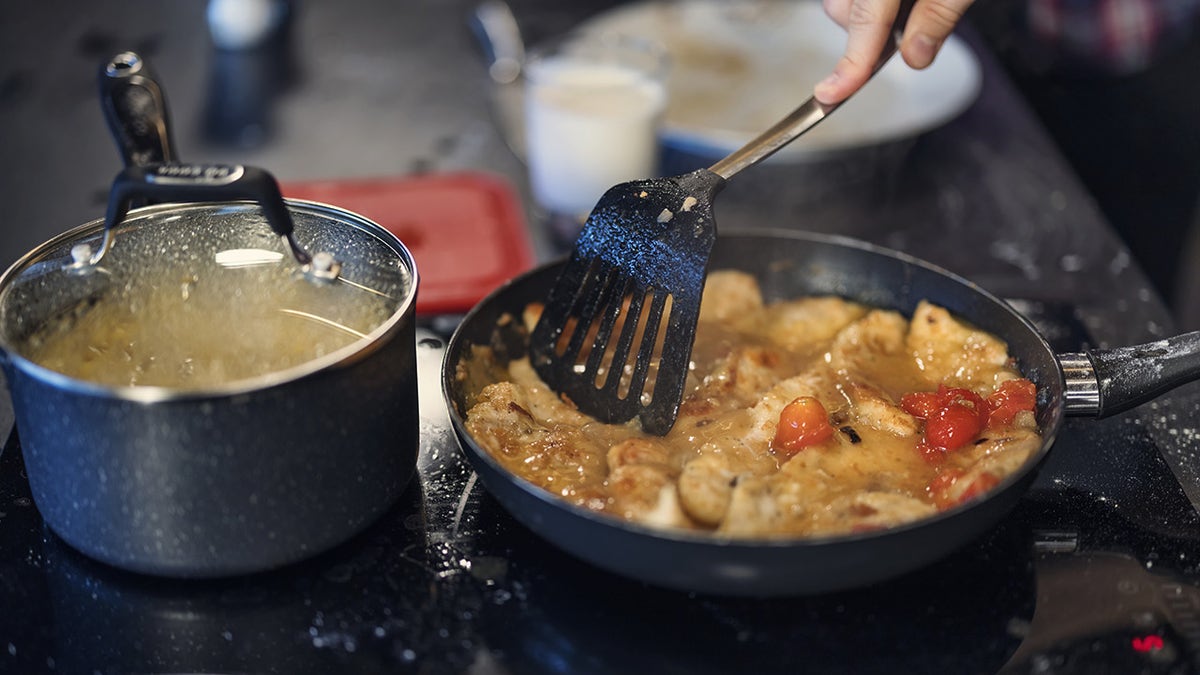 Person's hand seen using spatula to stir chicken and tomatoes simmering in a pa, as a pot of something else with cover on it sits on burner nearby.