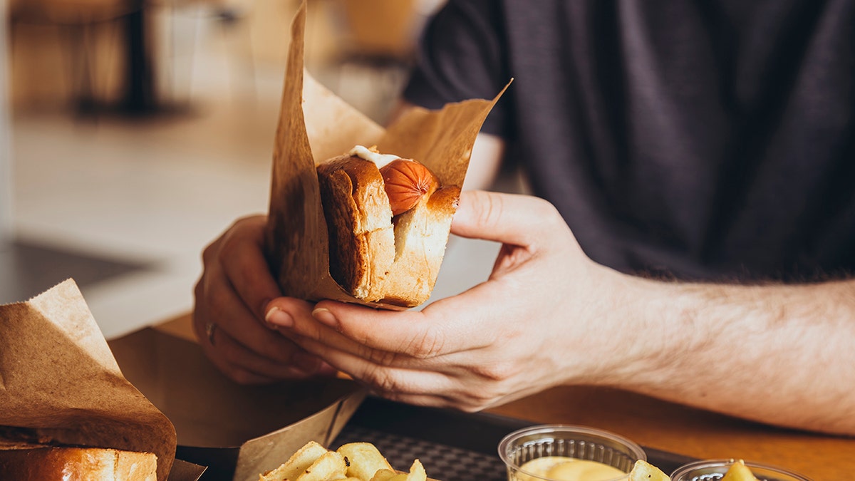 Man's hands seen up close as he holds hot dog in bun and paper. Men who ate highly processed foods gained more fat despite consuming the same calories as those who ate whole foods.