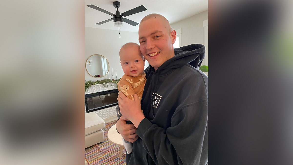Markus Schouten, pictured without hair, smiles while holding a baby in a living room decorated for the holidays.