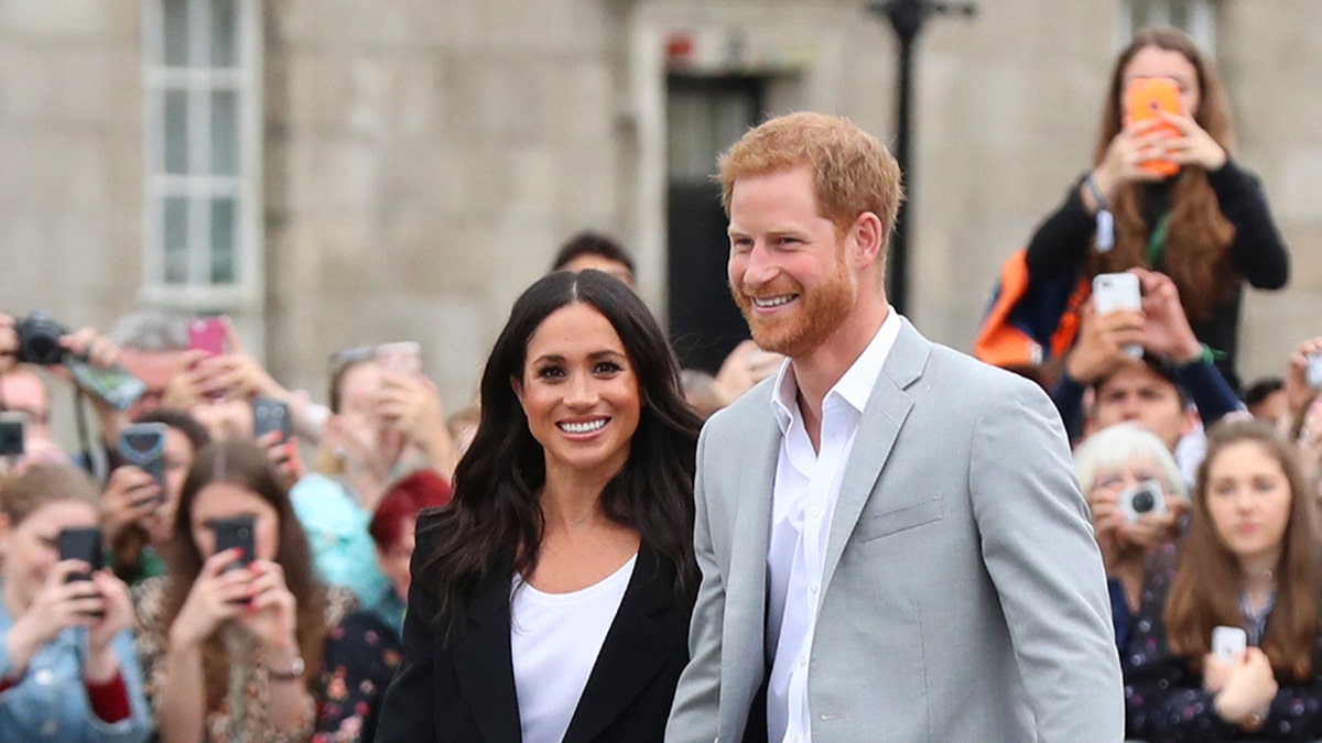 Prince Harry and Meghan Markle walking together and smile as a crowd takes their photo.