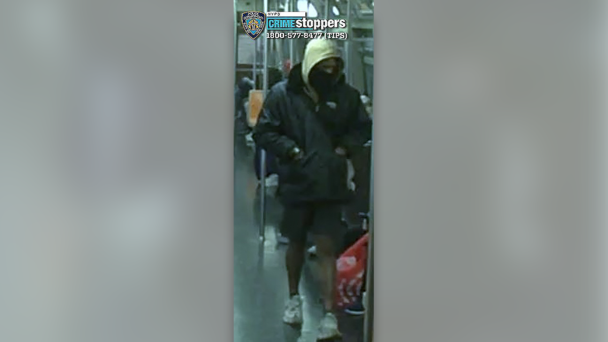 Surveillance image shows a man in a black jacket, with a tan hood up and a black mask, wearing shorts and white. sneakers, on a Subway car.