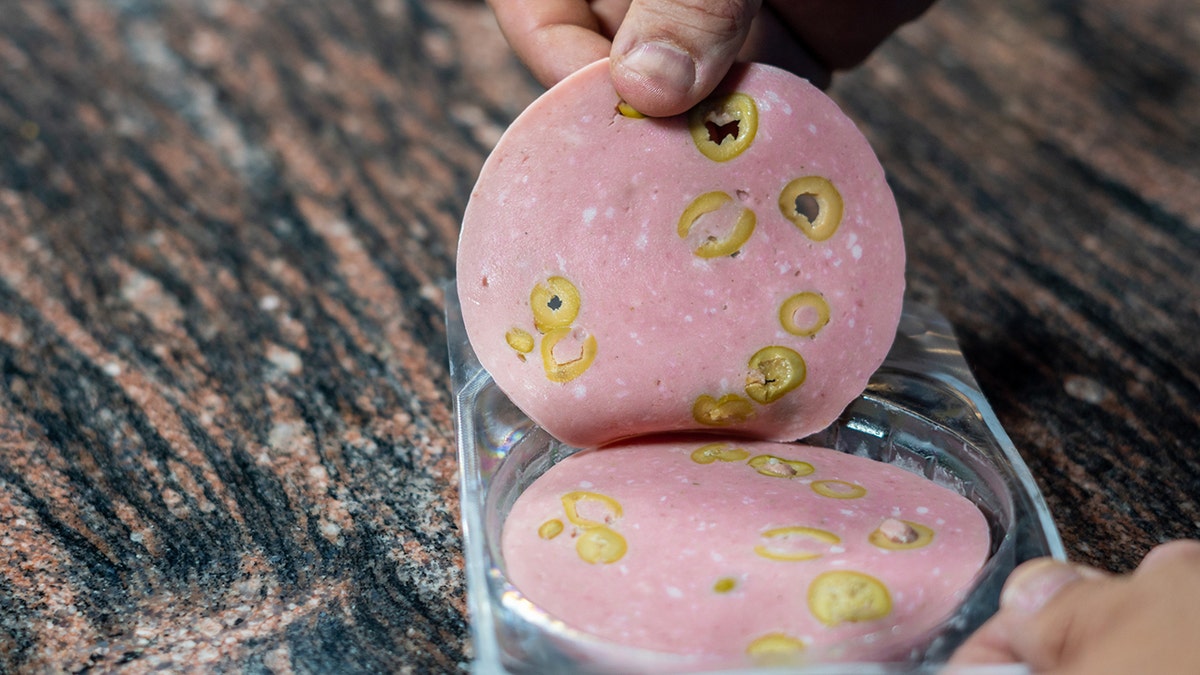 Person lifting slice of olive loaf lunch meat from container on counter