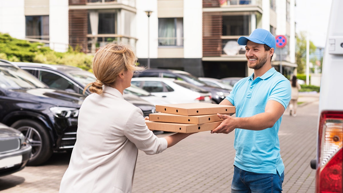 Smiling pizza delivery man wearing blue uniform delivers pizza boxes to a woman outside van in parking lot.