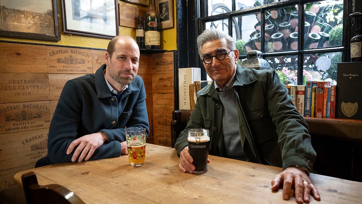 Prince William sitting next to Eugene Levy while they have beers at a pub.