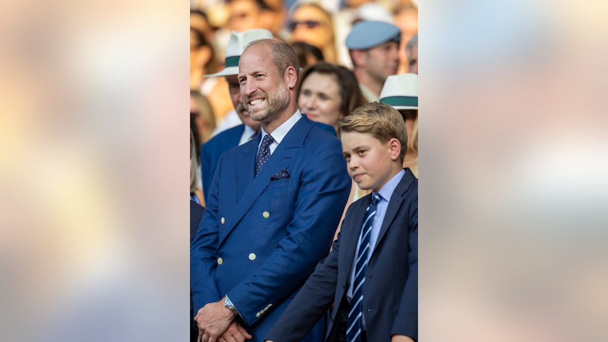 Prince William smiling in a suit next to his son Prince George as they admire a game with the crowd.