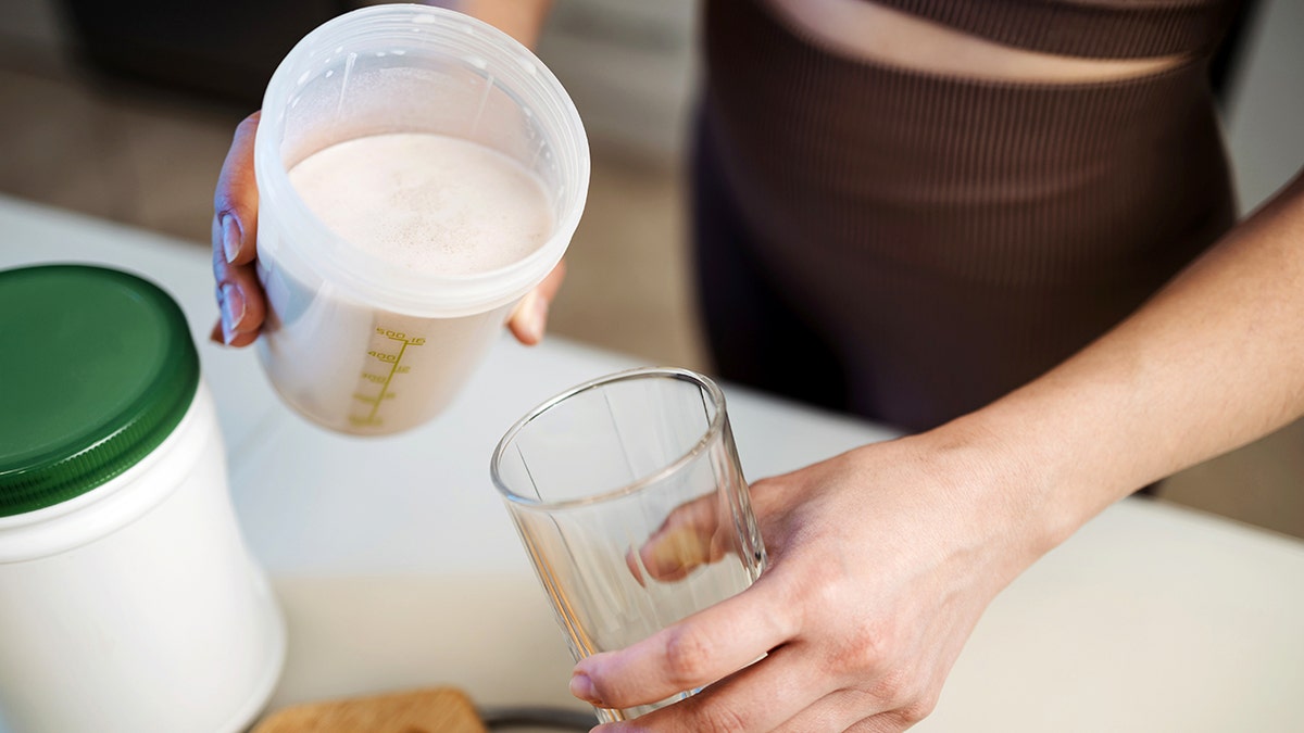 A woman holds a glass in one hand and a measuring cup filled with a smoothie.