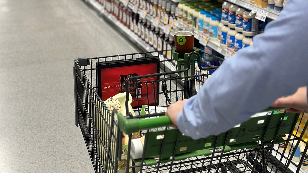 A Publix shopping cart is shown inside a supermarket with beer in a cup holder.