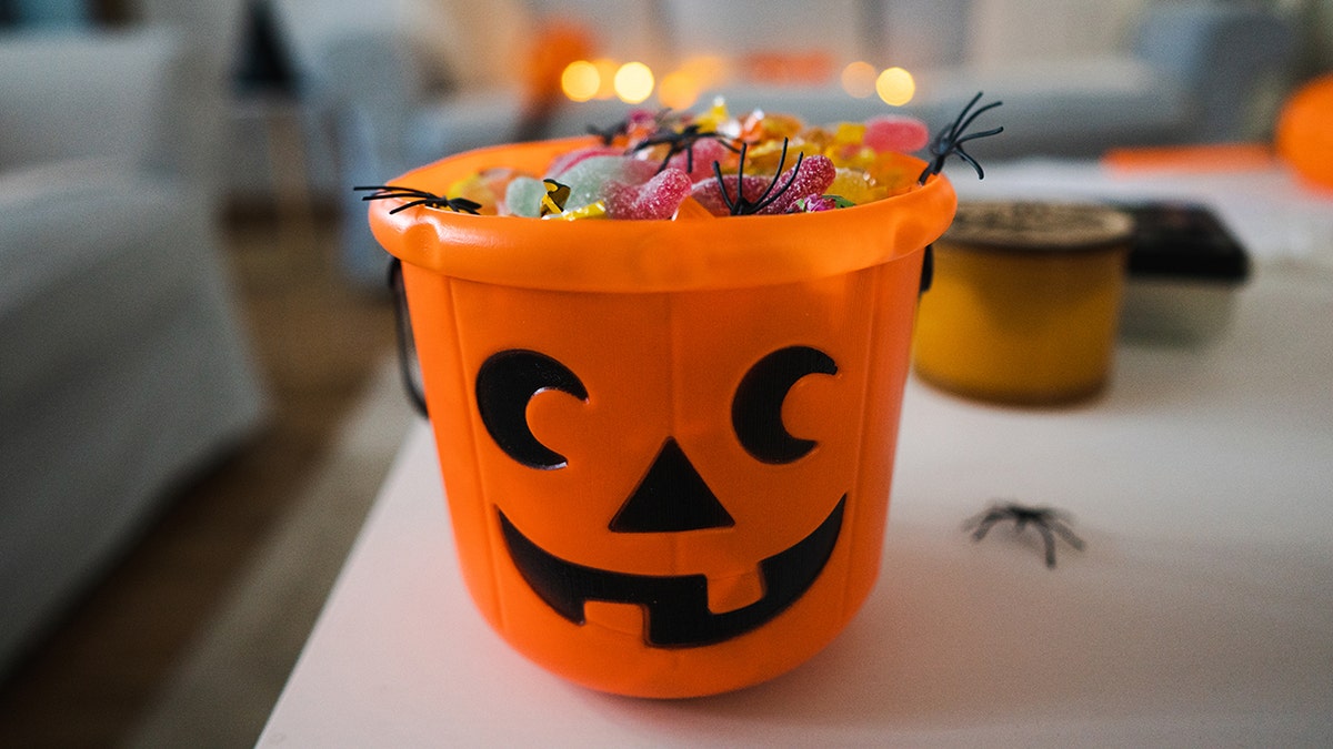 Plastic pumpkin bucket filled with candy, seen on counter with fake spiders scattered about.