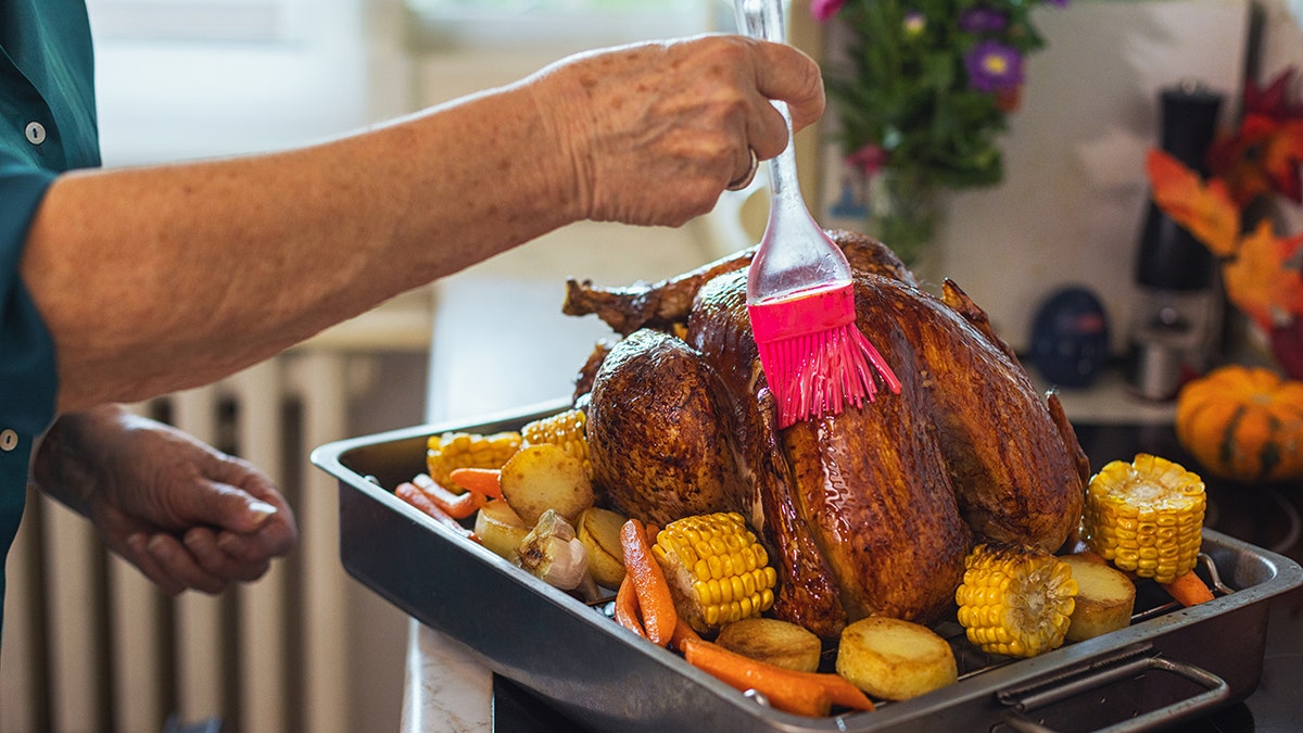 hands of woman seen brushing marinade on Turkey, prepared on a pan surrounded with veggies for roasting.