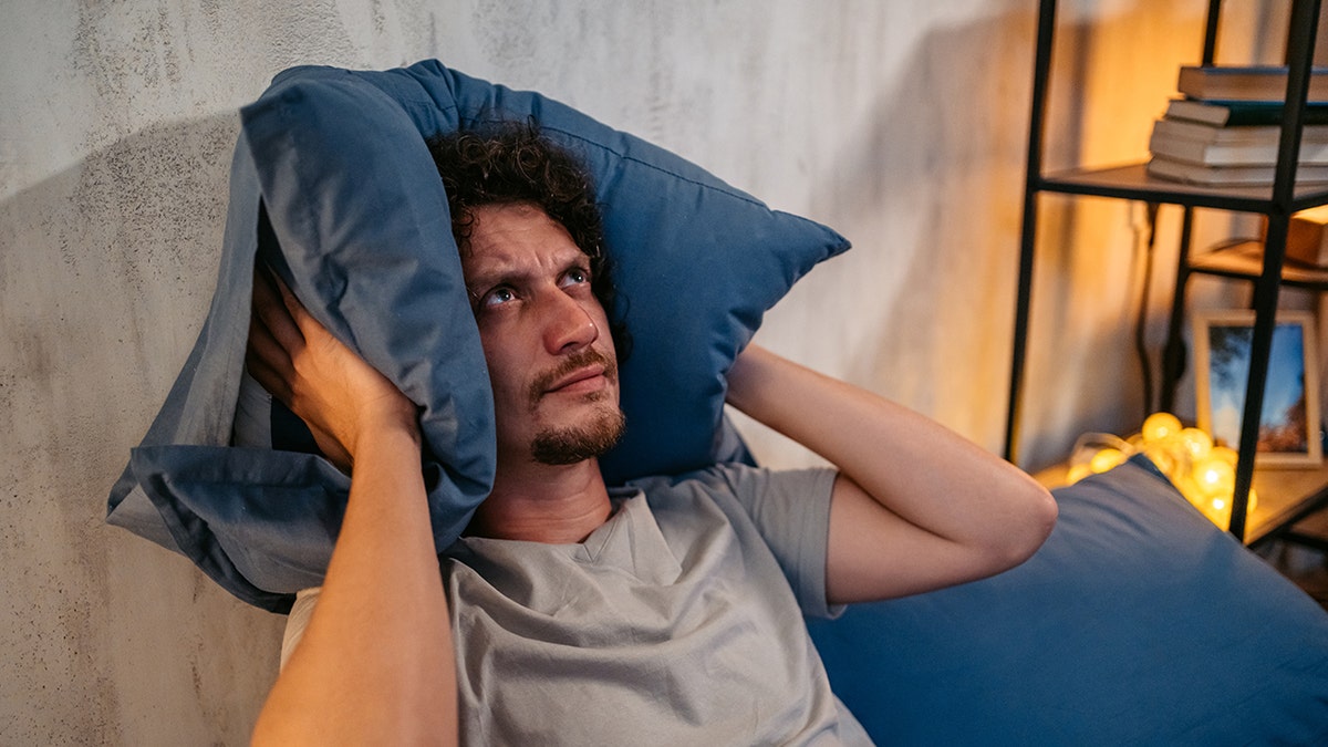 Man looking angry laying in bed holding pillow sides up to cover his ears, indicating loud noise disrupting his sleep.