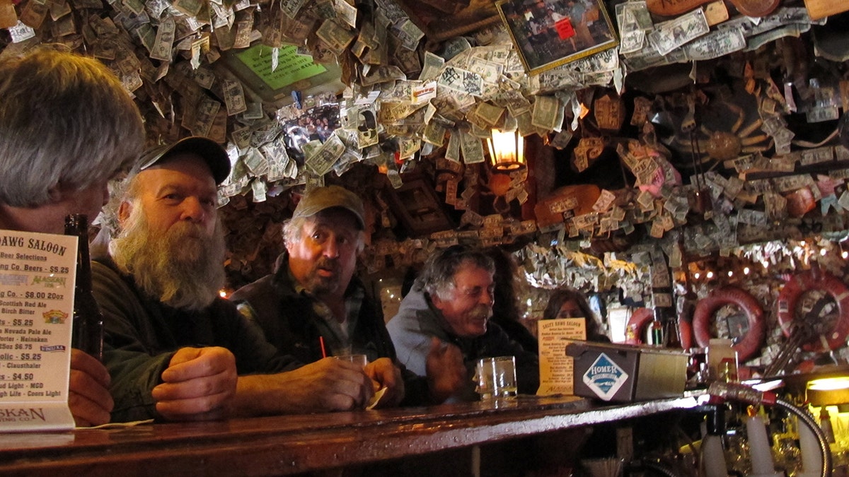 Homens sentados no Salty Dawg Saloon, no Alasca, notas de dólar e outras decorações revestindo as paredes atrás deles.