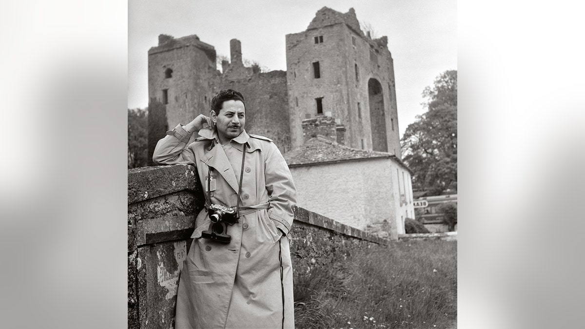 Sam Shaw wearing a trench coat in front of a castle in Europe.
