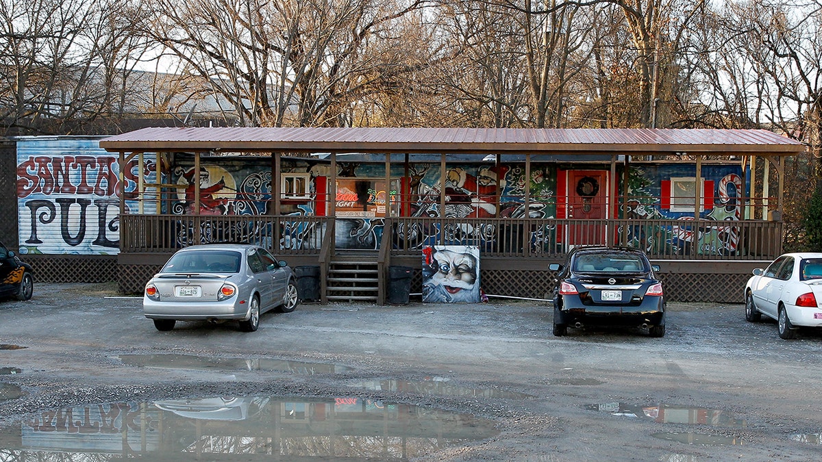 Do lado de fora do Santa's Pub em Nashville, Tennessee, localizado em um antigo trailer. Há carros estacionados do lado de fora, cercados por poças e árvores.