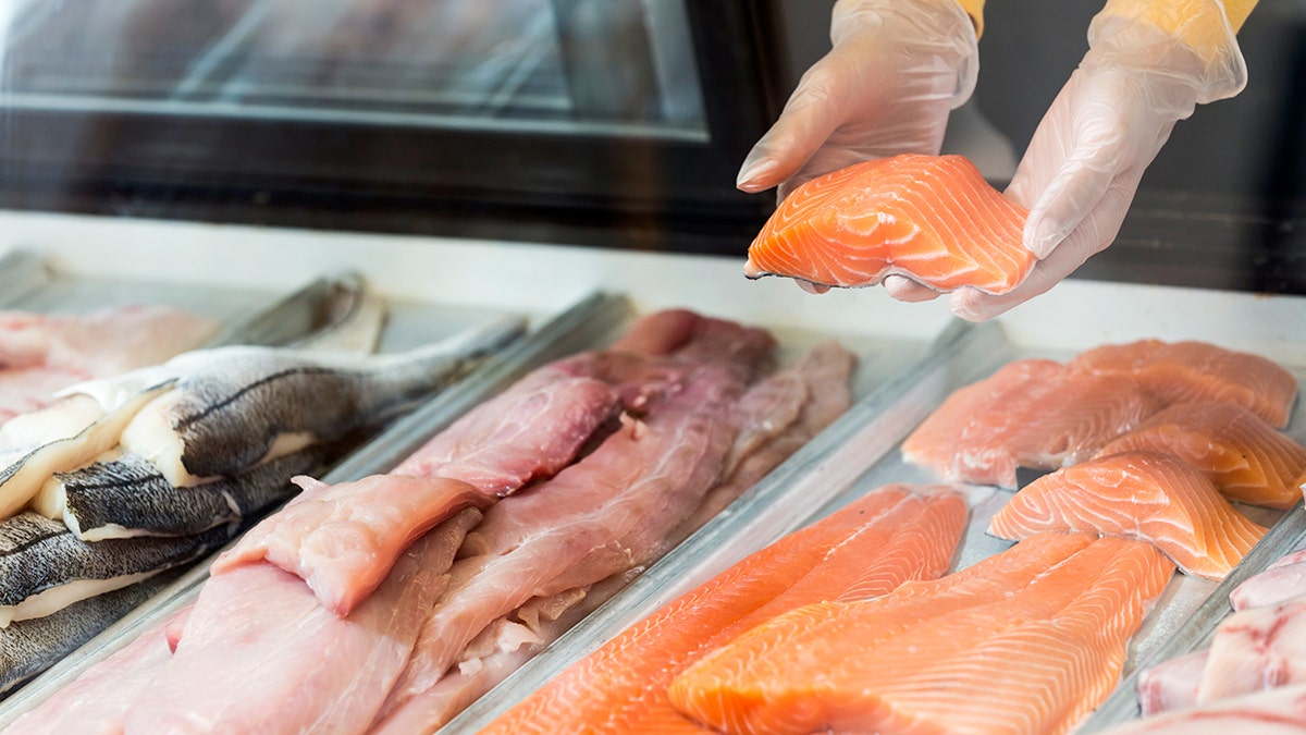 A grocery store employee's hands hold a piece of salmon inside a display case for seafood.