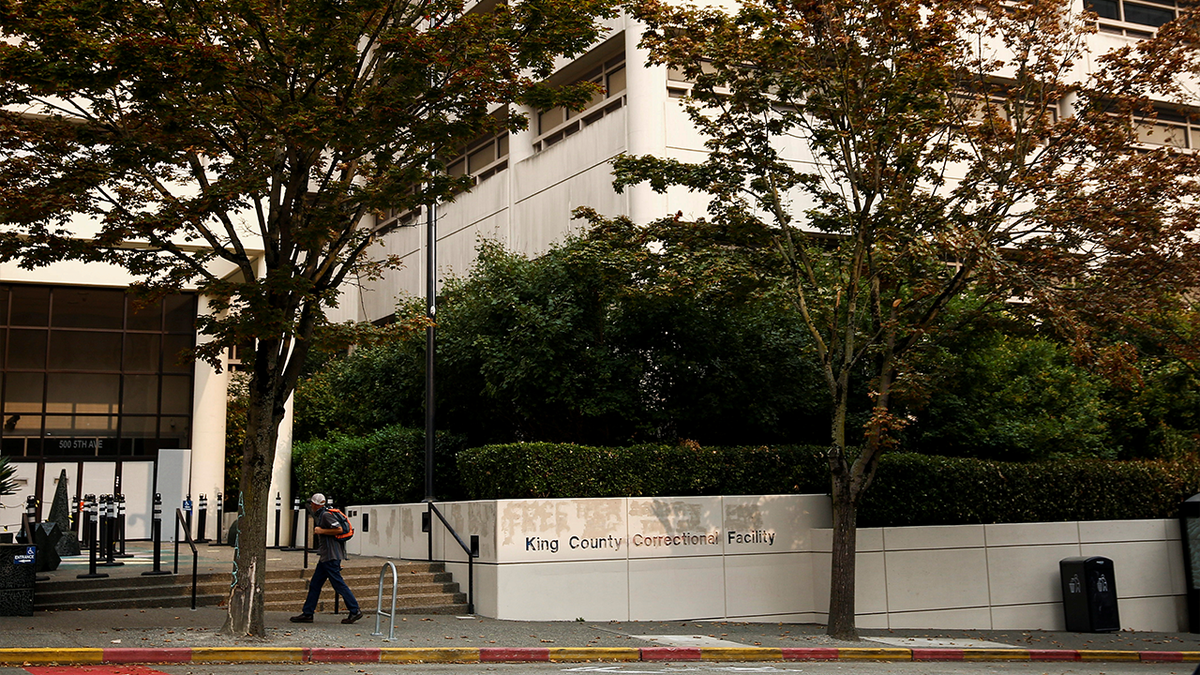 A pedestrian passes by the King County Correctional Facility in downtown Seattle, which has a row of trees and hedges in front of its white walls