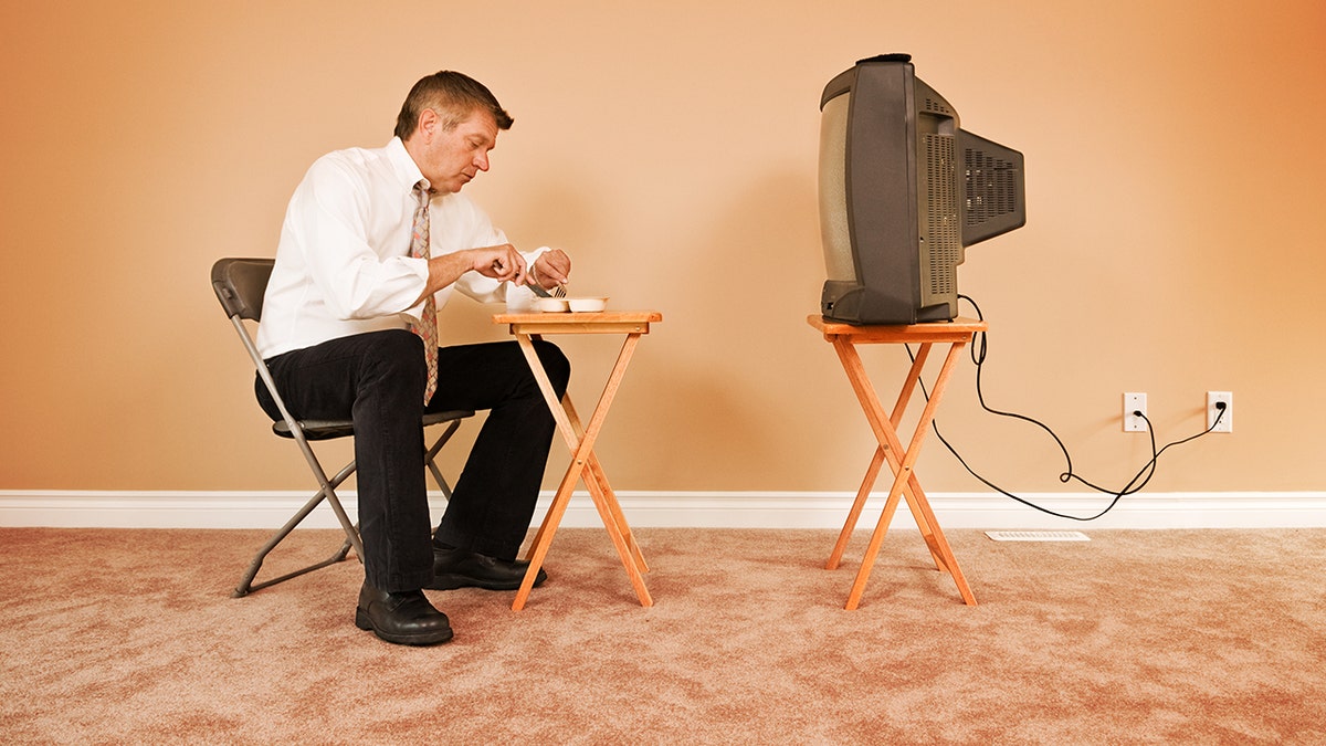 Man sitting at chair in front of folding table eating TV alone in front of television and empty living room