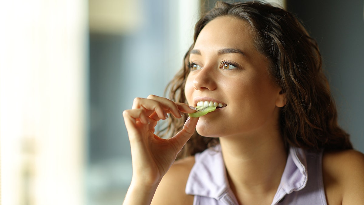 A woman looks outside while biting into a slice of kiwi at home.