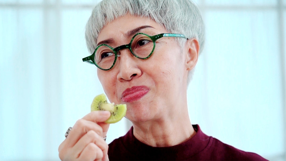 An Asian woman with gray hair and glasses holds a piece of kiwi in her hand after biting into it.
