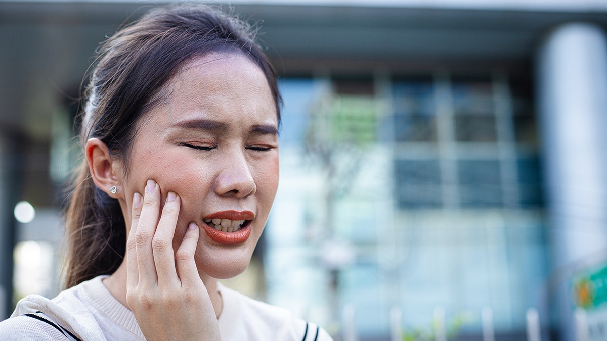 Young asian woman grimacing in pain, holding hand to jaw indicated tooth pain, gum disease, dental issues.