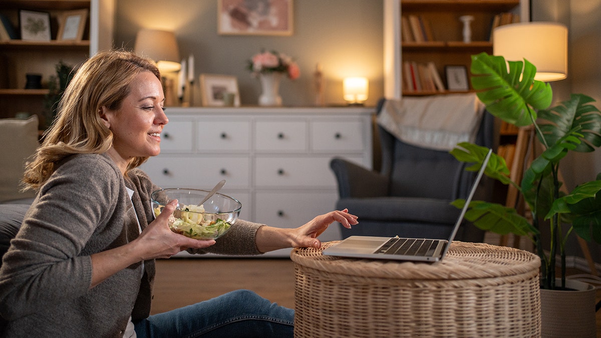 Woman sitting on floor eating salad with laptop on end table