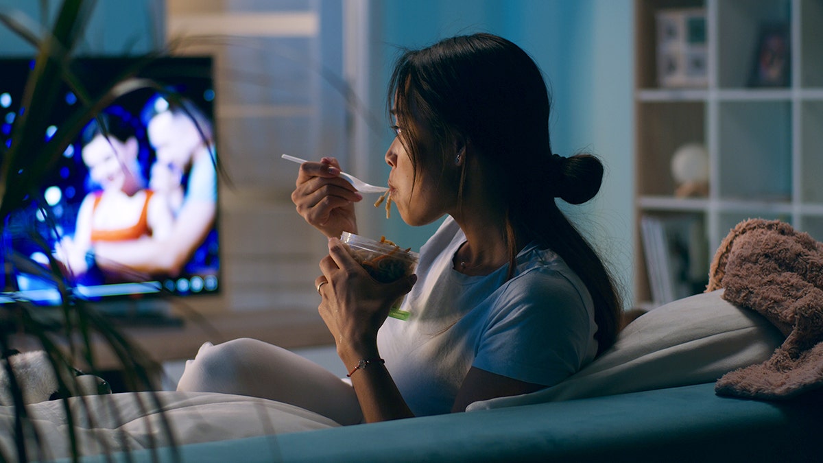 A woman watches TV on a couch while eating out of Tupperware with a plastic utensil.