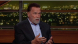 Arnold Schwarzenegger gestures while speaking during an interview on "Real Time with Bill Maher," with city lights glowing in the background.