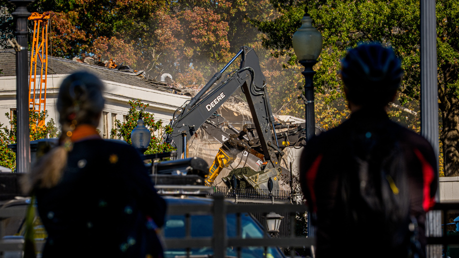 Pedestrians watch the facade of the East Wing of the White House being demolished
