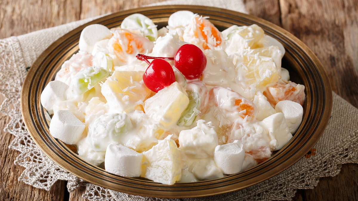 Ambrosia fruit salad with pineapple, oranges, grapes and coconut with marshmallow and vanilla yogurt close-up on a plate and doily.