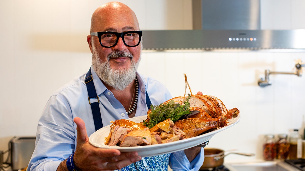Andrew Zimmern holds a plate of meat in a kitchen.