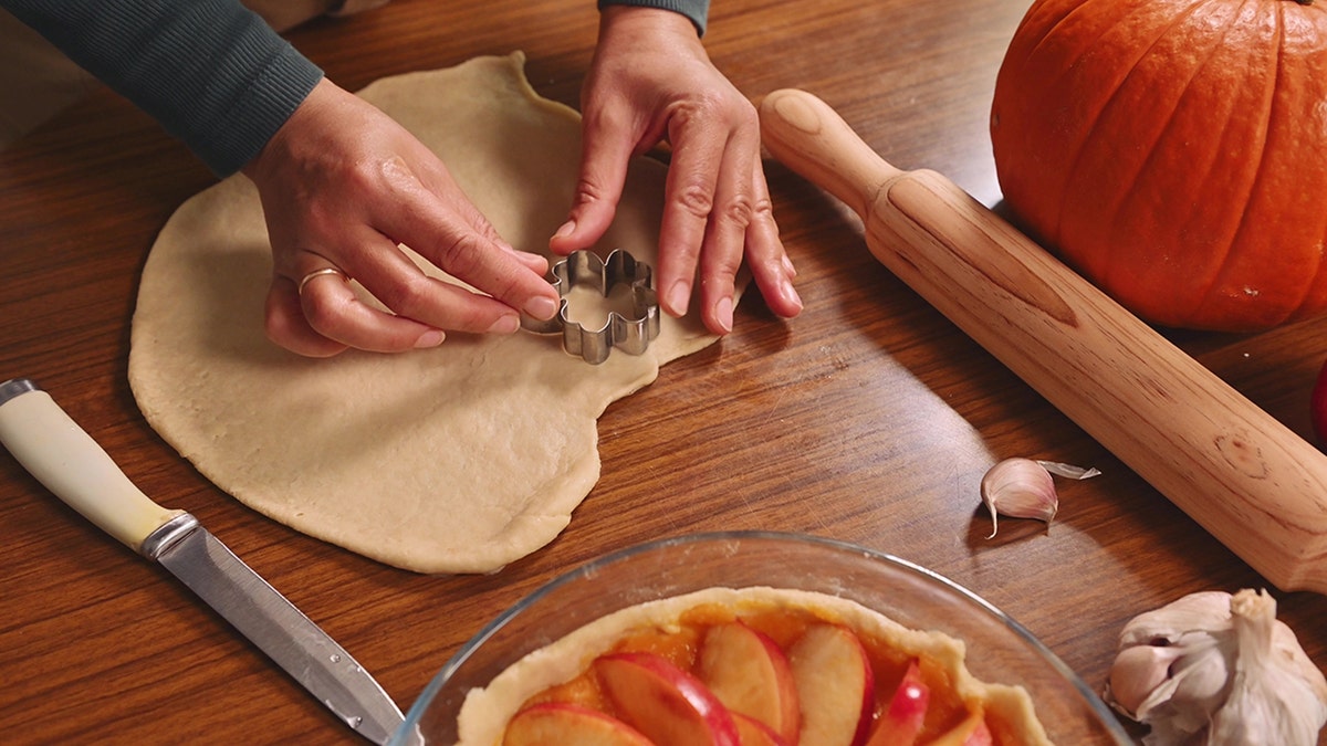 Woman's hands using a cookie cutter on dough, surrounded by pumpkins, rolling pin, and ingredients, making an apple pie on a wooden table.