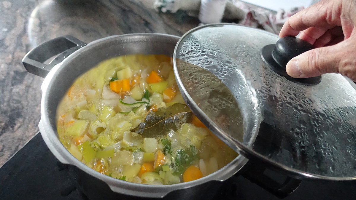 Person's hand seen removing lid from pot of soup on stove, bay leaf at center of pot.