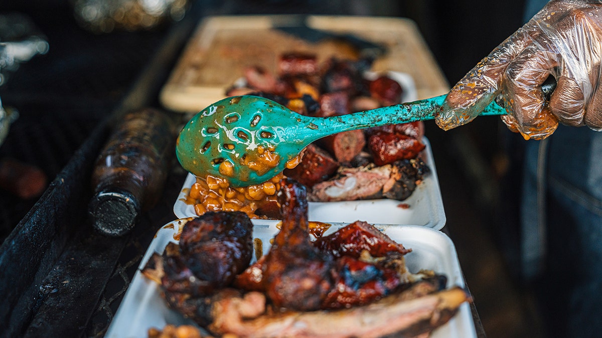 Chef's hand serving baked beans onto a takeout container of BBQ meat in Texas.