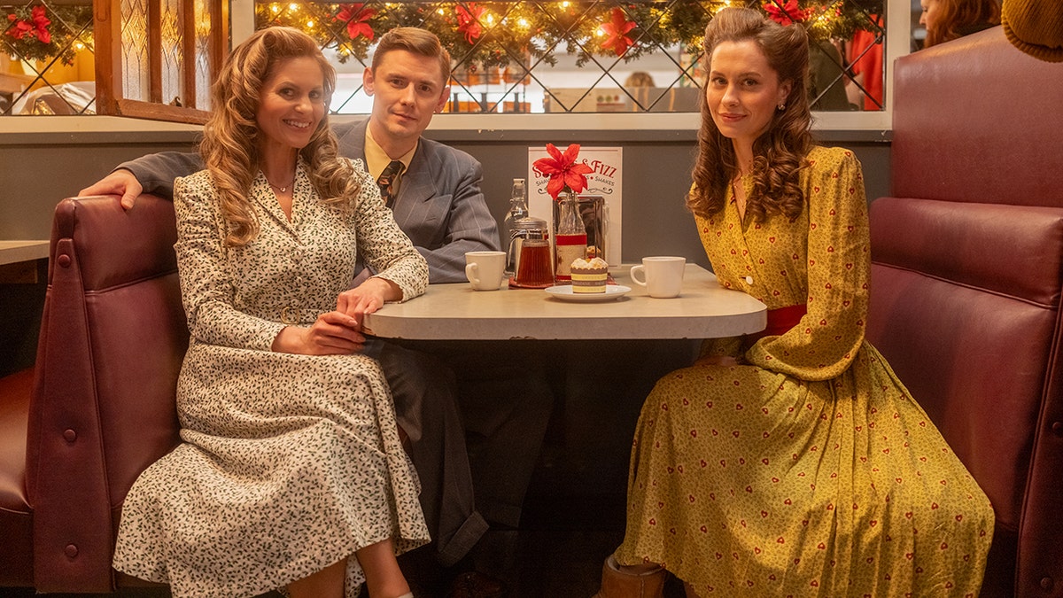 Candace Cameron Bure, Natasha Perry and Paul Greene member dressed in vintage-style clothing sit together at a diner booth decorated for the holidays.