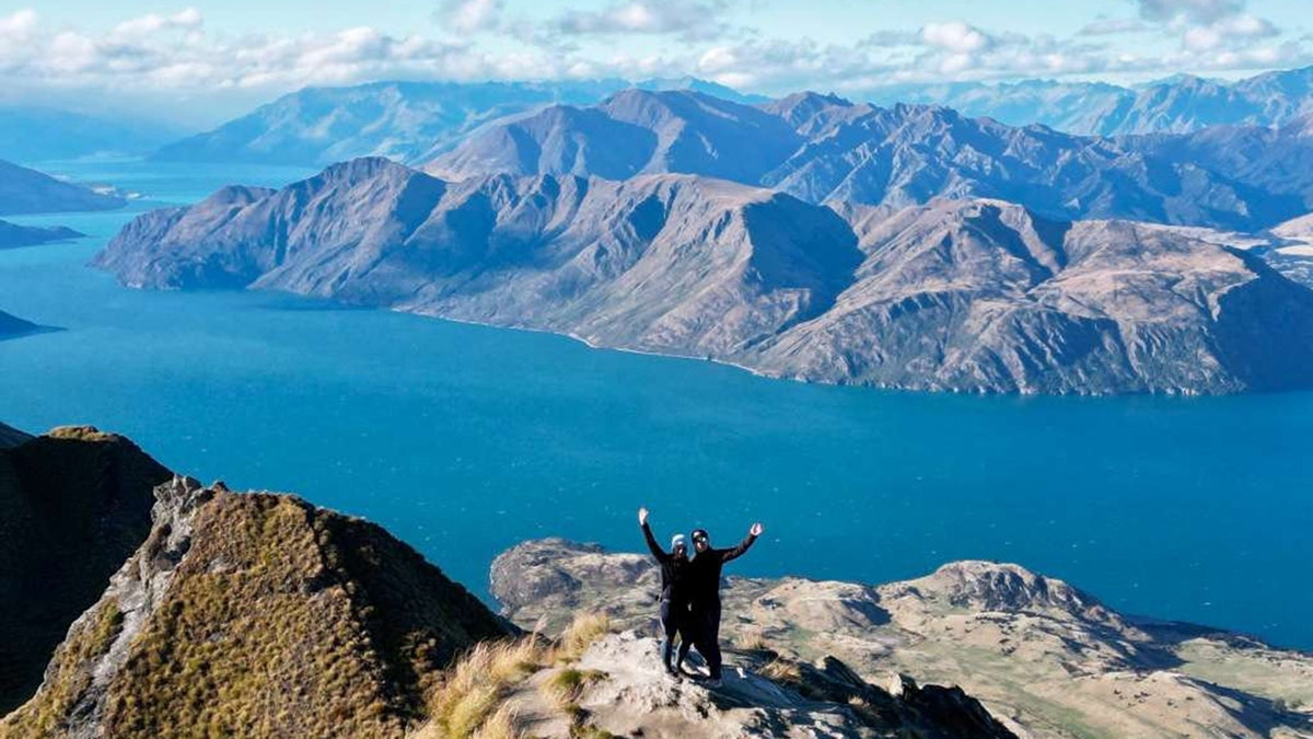 Two hikers stand triumphantly on a mountain ridge overlooking a deep blue lake and rugged mountain range