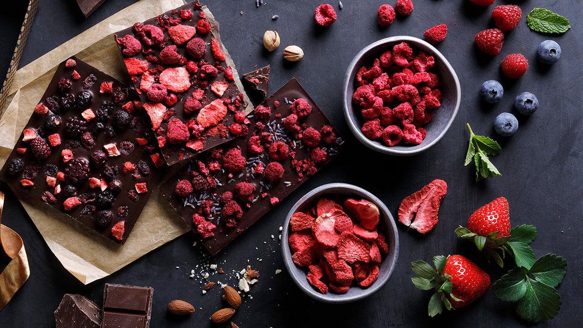 Dark chocolate bark seen topped with dried berries, nuts, sea salt, mint and other toppings scattered about.