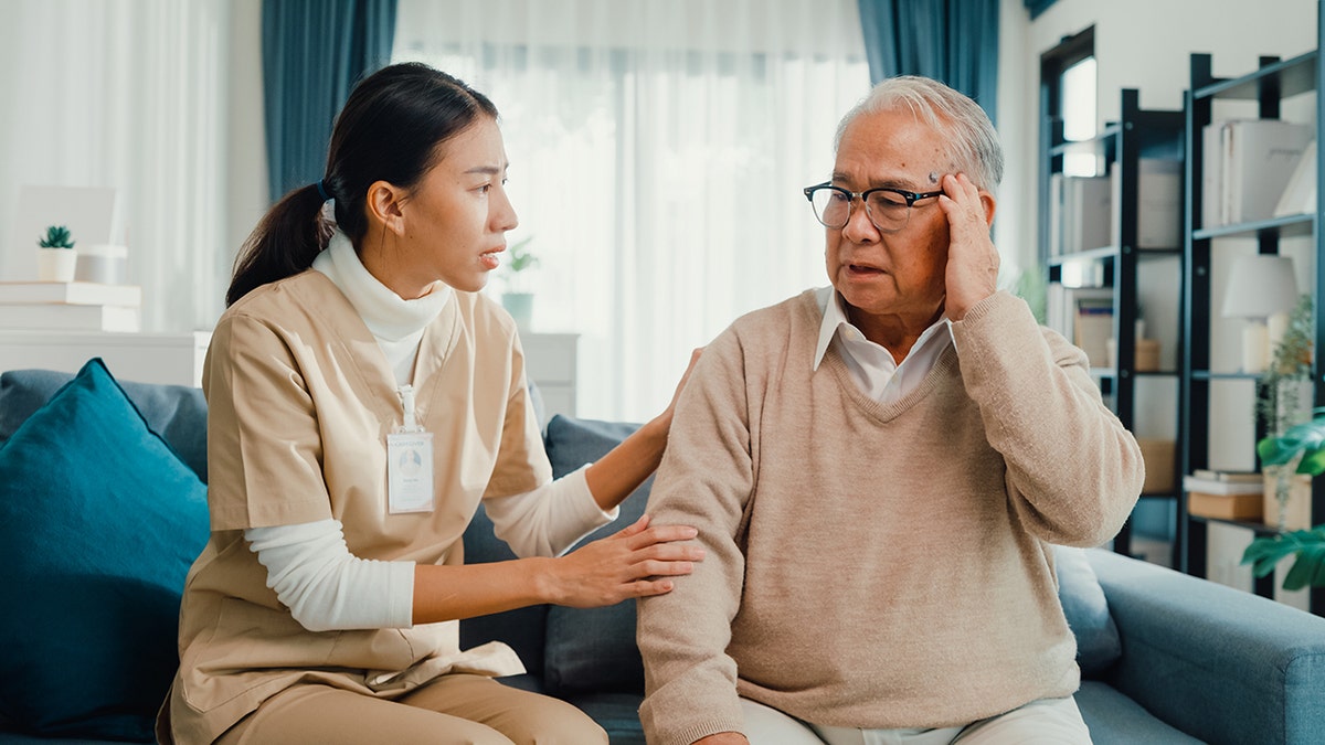 A caregiver speaks with concern to an older man holding his head, sitting on a couch.