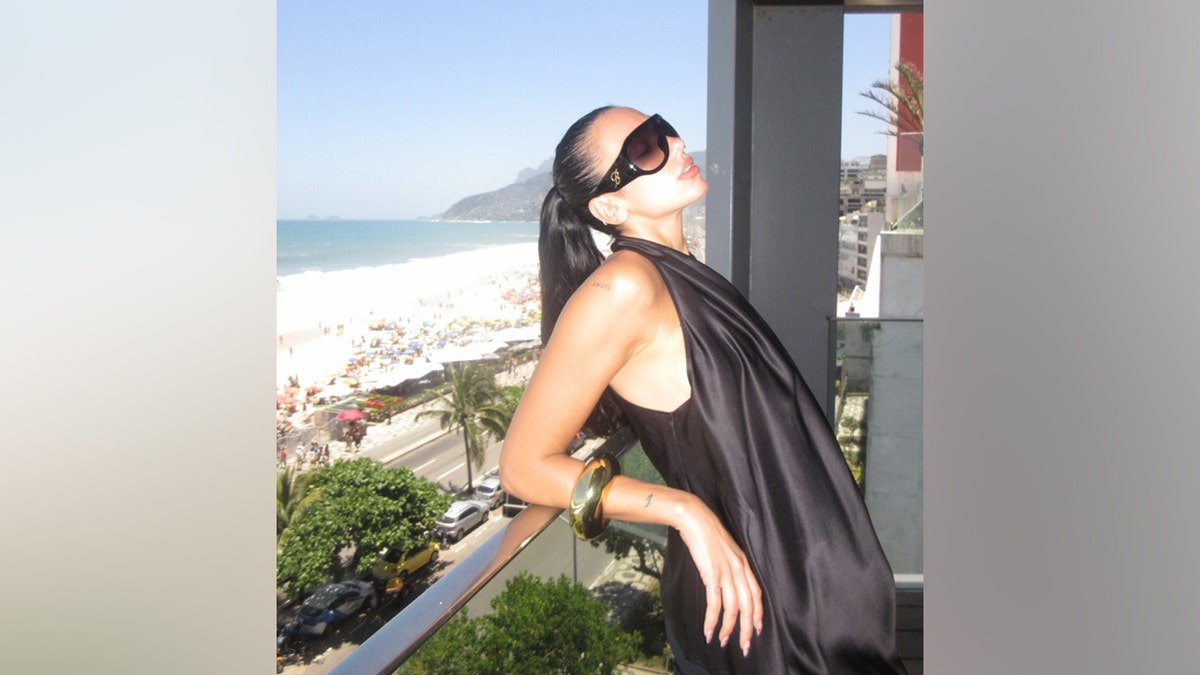 Dua Lipa posing on a balcony in Brazil, wearing oversized sunglasses and a black outfit with the beach in the background.