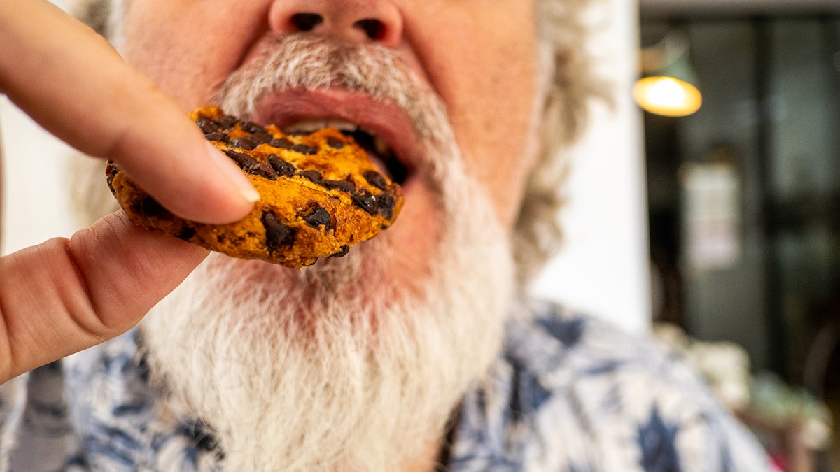A bearded man eats a chocolate chip cookie.