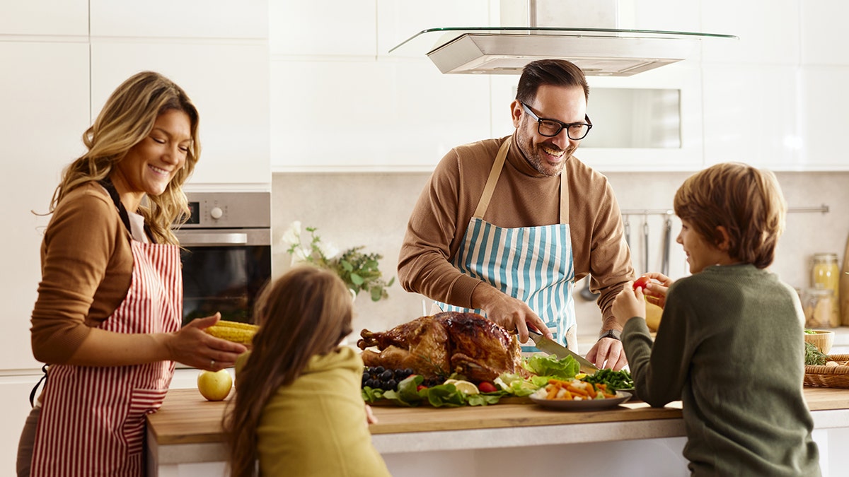 A family preparing a meal together in a bright kitchen. A man in a striped apron carves a roasted turkey while a woman and two children smile and help with the food.