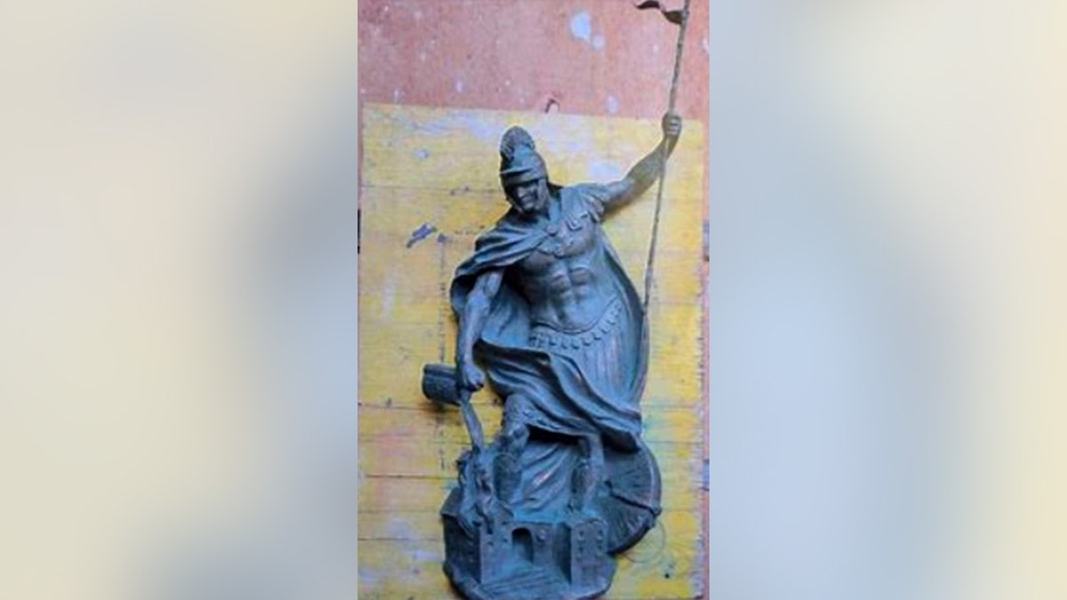 A detailed bronze sculpture of Saint Florian dressed in Roman armor, holding a vessel and pouring water onto a small burning structure beneath him.