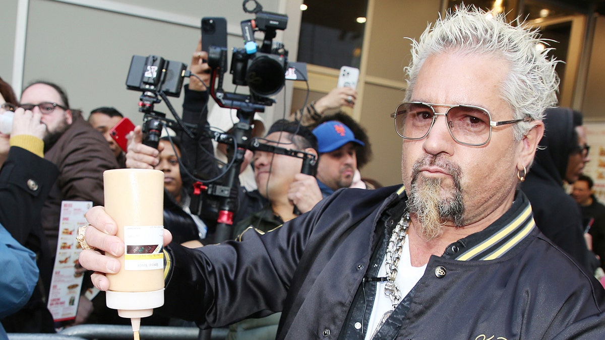 Guy Fieri holds a squeeze bottle while standing in front of a crowd of onlookers and cameras.