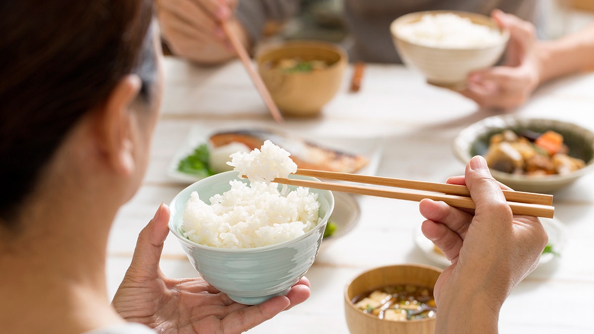 An Asian woman holds a clump of white rice with her chopsticks.