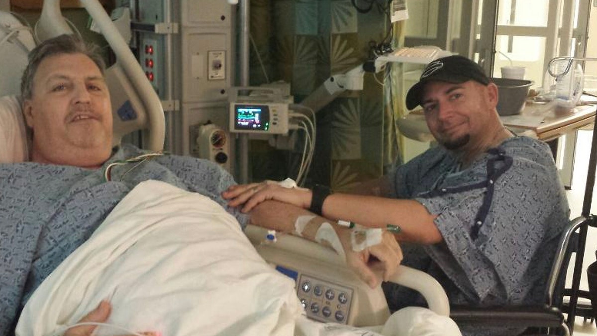 One man is lying in a hospital bed, while another man is sitting next to him in a wheelchair.