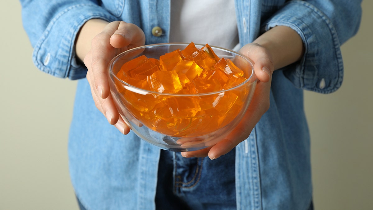 Woman in denim jeans and denim shirt holding glass bowl of orange Jell-O cubes.