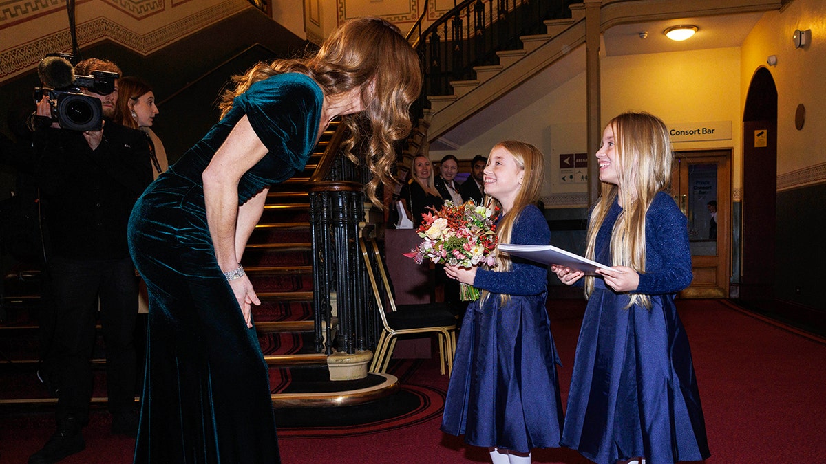 Kate Middleton bending down to greet two smiling twin girls wearing matching blue dresses.