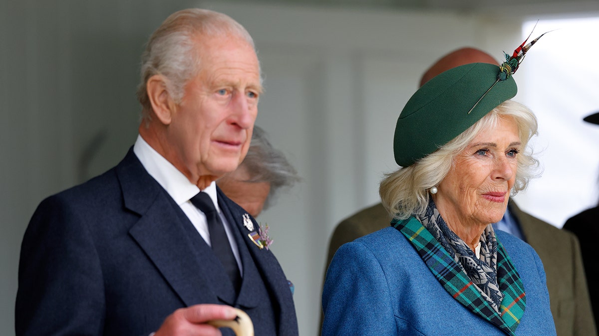 King Charles in a dark blue suit standing next to Queen Camilla as they both watch on.