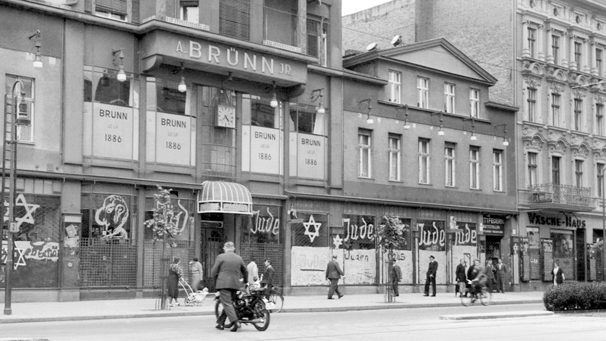 A shattered storefront displays signs of targeted destruction amid rising persecution in Nazi Germany.