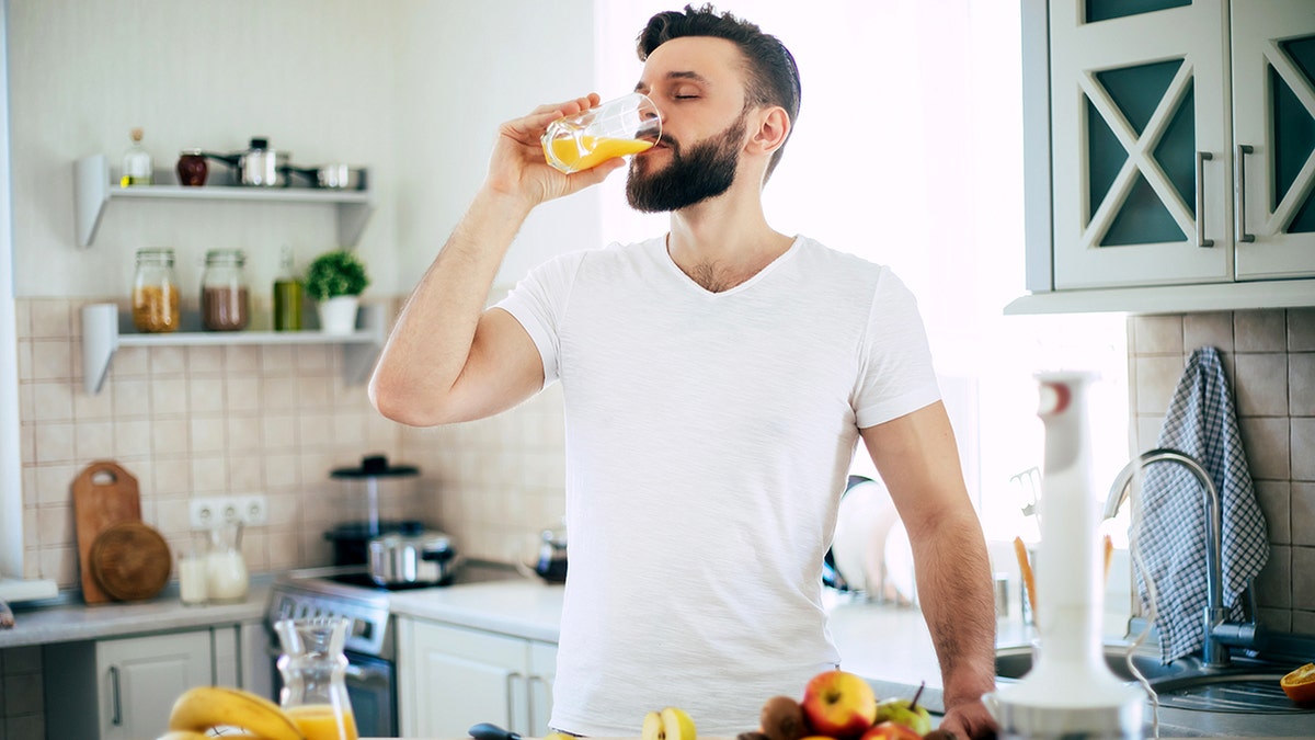 A man wearing a white undershirt drinks orange juice from a glass with his eyes closed.